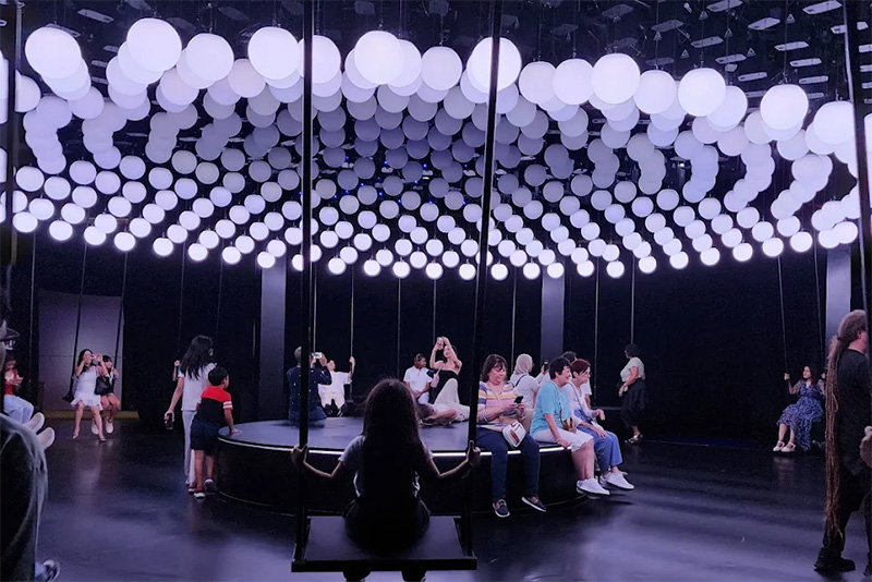 A large, dark room filled with hundreds of glowing white balloons suspended from the ceiling, with people sitting and relaxing on the floor beneath the installation at the Balloon Museum