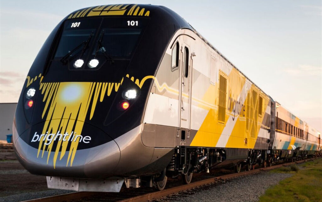 A sleek, modern Brightline high-speed train with a bright yellow and white exterior, traveling along the Florida rail corridor under a clear sky.