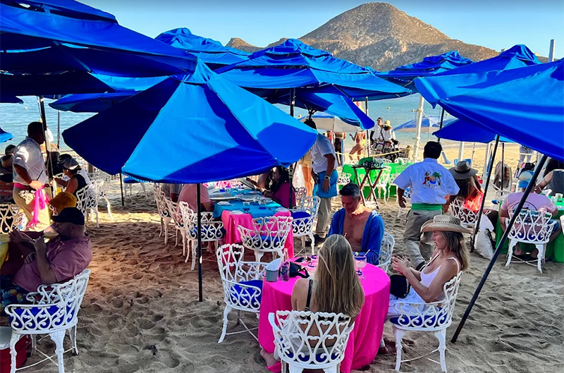 A crowded open-air beachfront restaurant, The Office on Médano Beach in Cabo San Lucas, with people sitting at blue and pink-clothed tables with umbrellas in the sand near the water and a clear blue sky.
