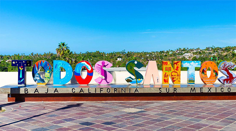 A colorful and large "Todos Santos" sign made of individual block letters with vibrant patterns, set against a backdrop of a sunny plaza with palm trees and a bright blue sky in Baja California Sur.