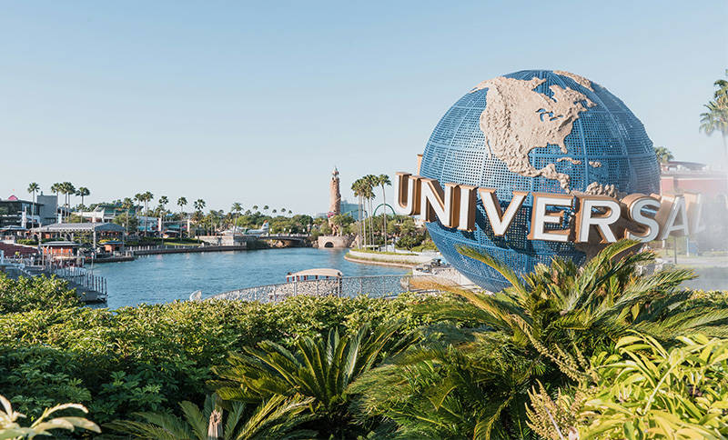 A wide daytime shot of the iconic Universal Studios Florida entrance plaza, featuring the large rotating "Universal" globe in front of the ornate archway and palm trees under a clear blue sky.