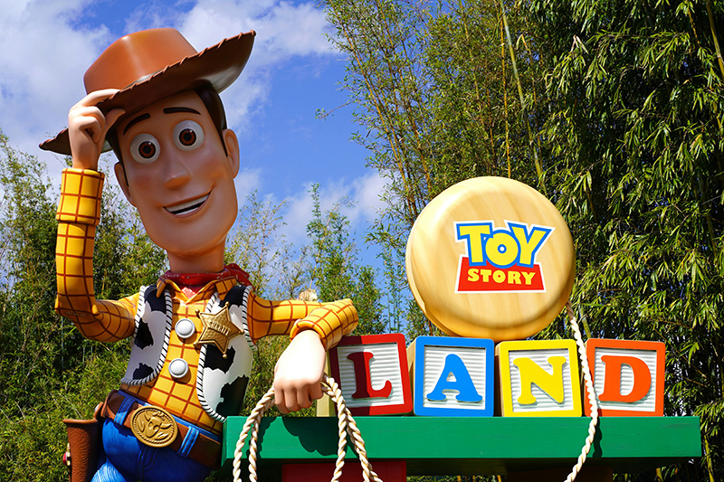 Woody leaning against the Toy Story Land entrance sign with the Slinky Dog Dash coaster in the background under a blue sky