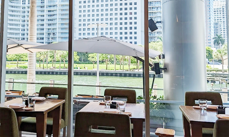 Interior of Zuma Miami featuring modern wooden tables, floor-to-ceiling windows with views of the Miami skyline and river, and a sophisticated, neutral-toned dining room