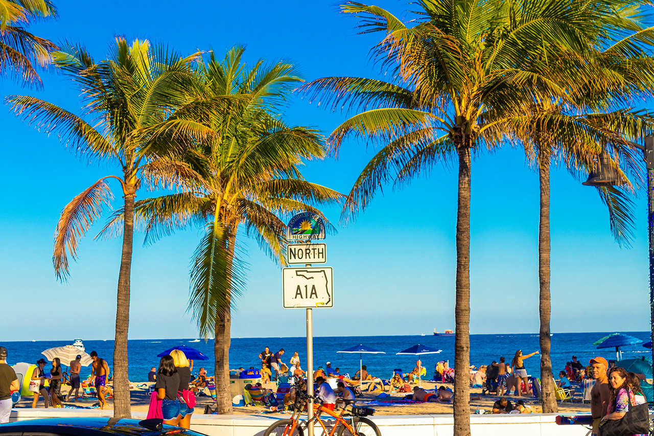 A serene coastal view of a Florida beach featuring soft white sand in the foreground and clear turquoise water stretching toward the horizon under a bright, clear sky.
