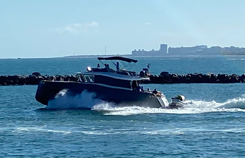 A close-up view of a large, dark-hulled motor yacht cruising through the choppy, deep blue waters of the bay, with the distant Miami skyline and low-lying islands visible on the horizon under a soft, hazy sky.