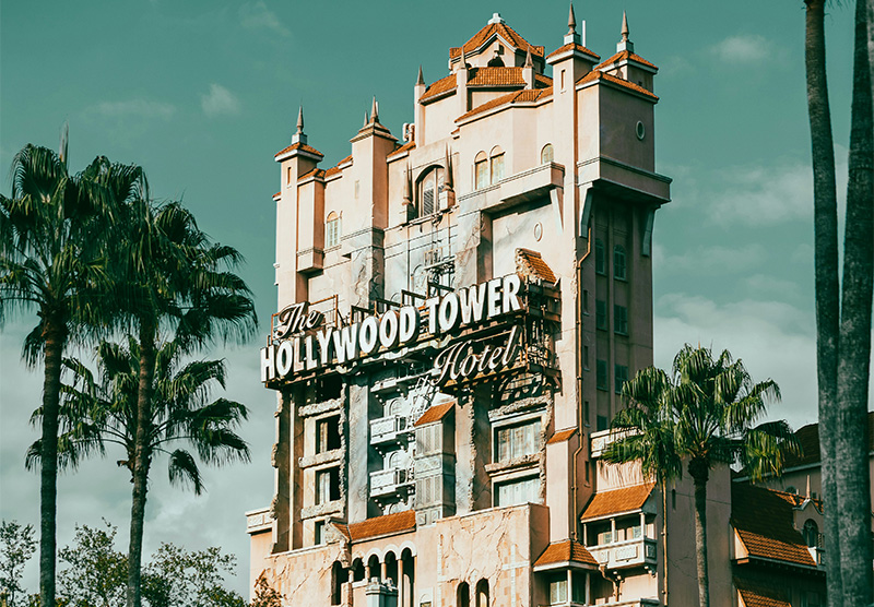 A tall, weathered, Art Deco-style hotel with a "Hollywood Tower Hotel" sign and scorched markings on the facade, framed by palm trees and a bright sky.