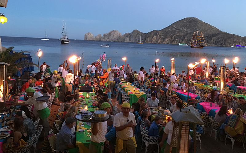A lively scene at a beach club on Médano Beach in Cabo San Lucas, showing travelers enjoying drinks at tables set directly in the sand with the turquoise ocean and Land's End rock formations in the background.