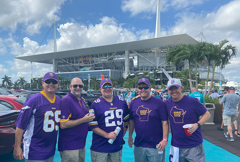 A group of fans wearing purple and gold Minnesota Vikings jerseys and gear standing in the seating bowl of Hard Rock Stadium during a sunny game day.