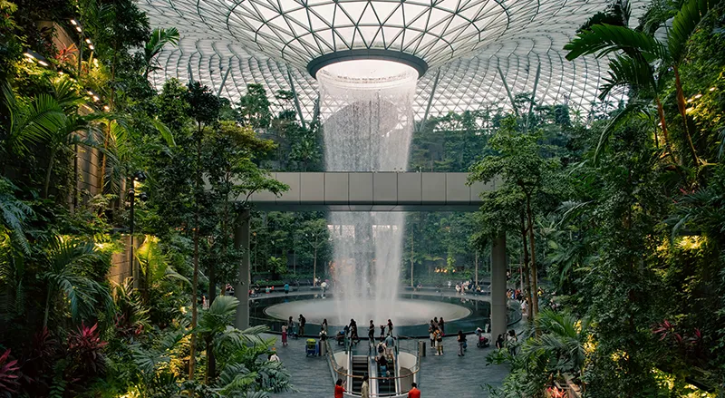 Indoor rain vortex waterfall cascading through the center of the lush, glass-domed Jewel Changi Airport in Singapore.