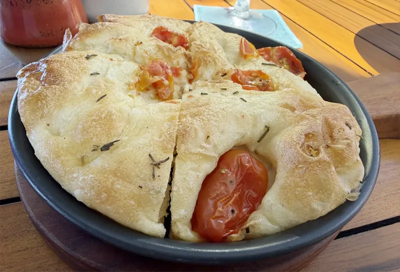 Freshly baked focaccia bread topped with cherry tomatoes, olives, and fresh herbs, baked to a golden brown on a wooden board.