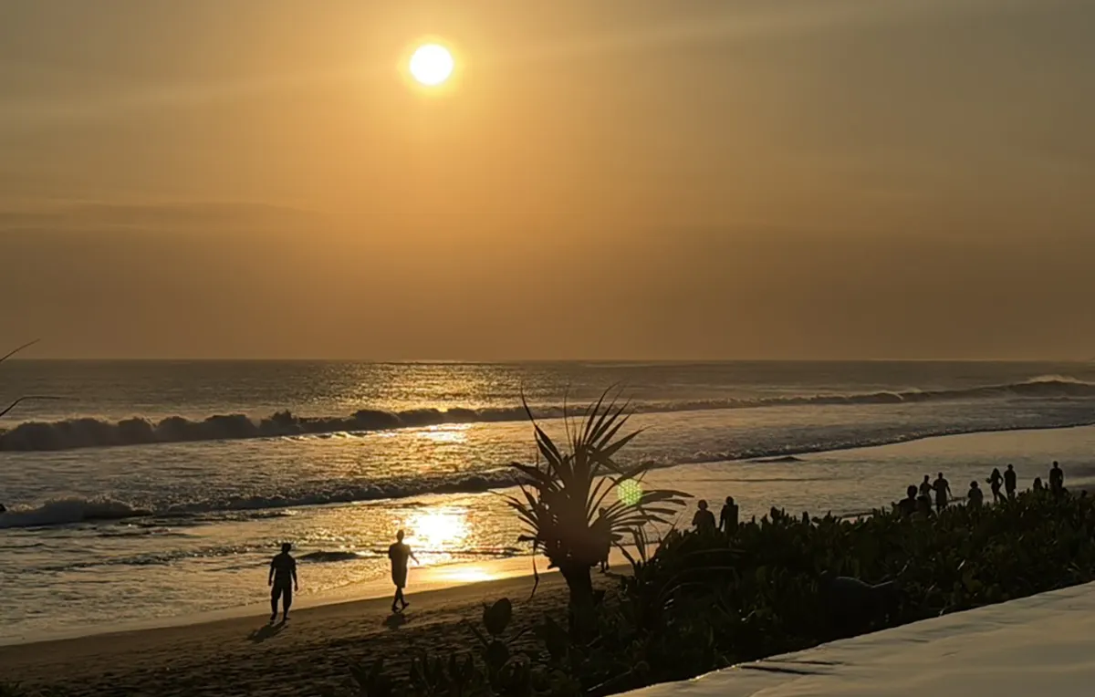 Golden sunset over a wide Bali beach with waves washing ashore and silhouetted figures walking along the wet sand.
