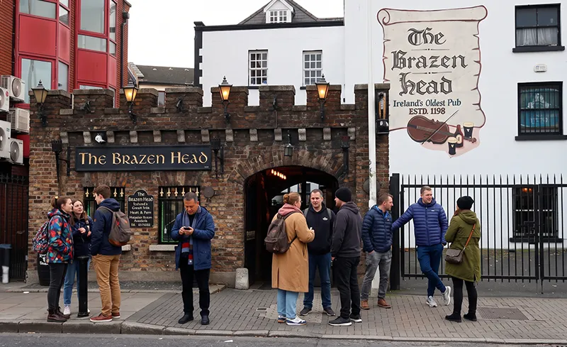 A natural, casual crowd of tourists gathered on the sidewalk outside the historic brick and stone exterior of The Brazen Head pub in Dublin, Ireland, illustrating the busy visitor crowds that critics worry AI travel chatbots will continue to concentrate at the same famous spots.