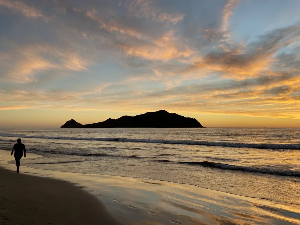 Lone beach walker beside the surf at sunset with an island silhouetted offshore in Mazatlán.