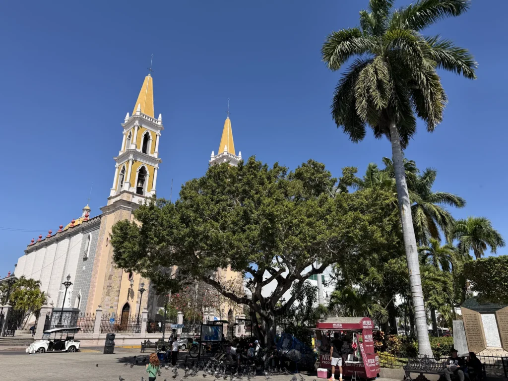 Yellow cathedral towers rising above trees against a blue sky in Mazatlán.