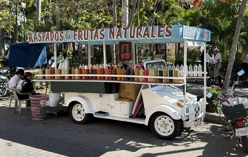 White raspado cart in Mazatlán’s Centro with colorful syrup bottles beneath a Raspados de Frutas Naturales sign.
