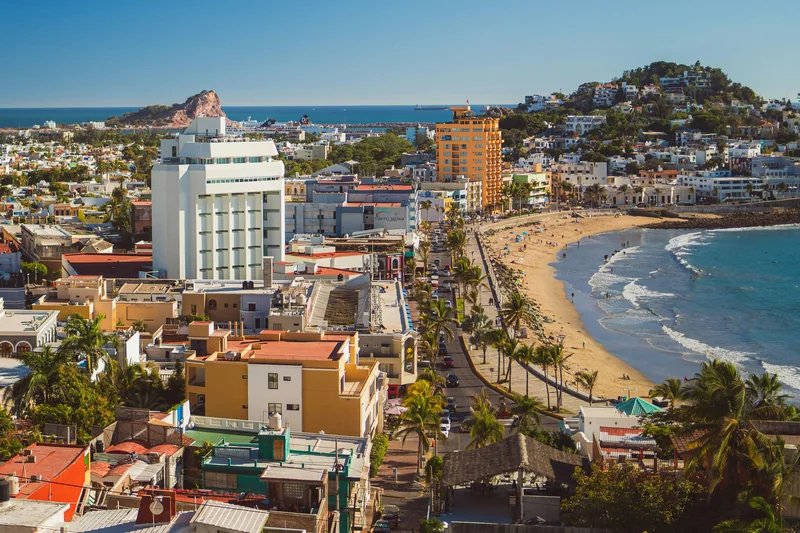 Elevated view of Mazatlán’s waterfront, with colorful buildings, palm trees, a curved beach, and the Pacific Ocean.