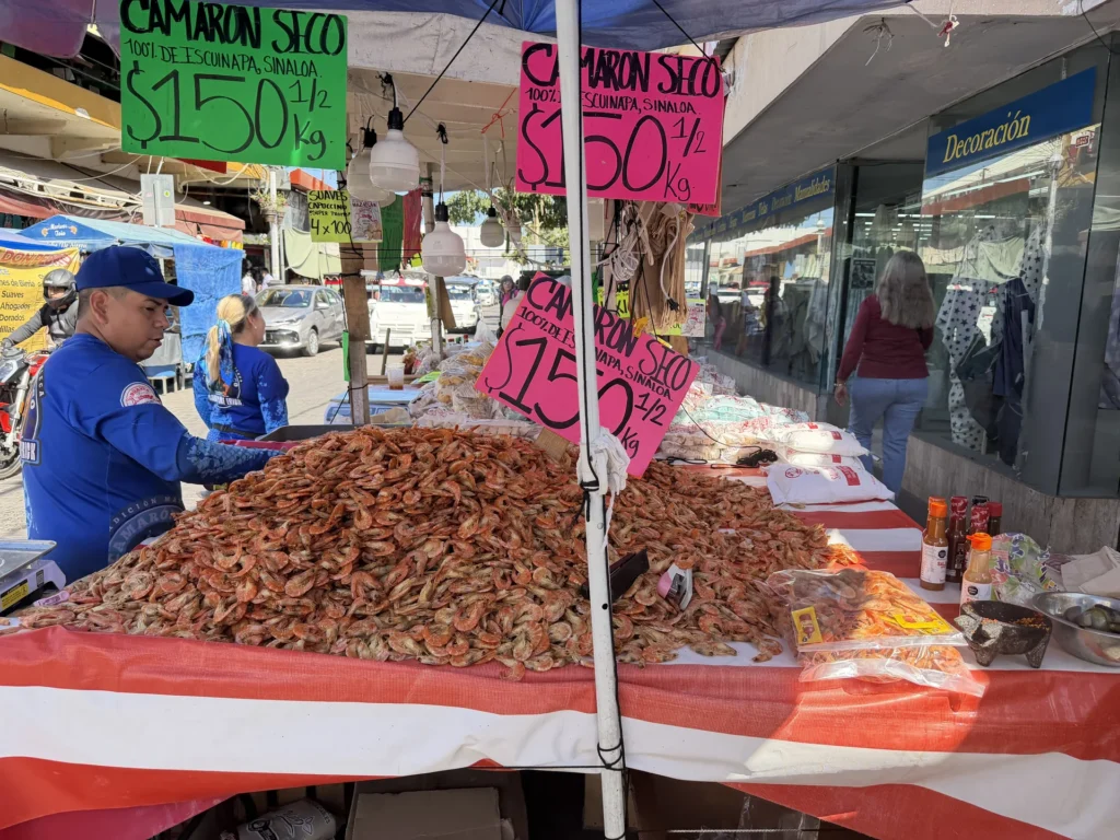 Market vendor behind piles of dried shrimp with signs advertising camarón seco in Mazatlán.