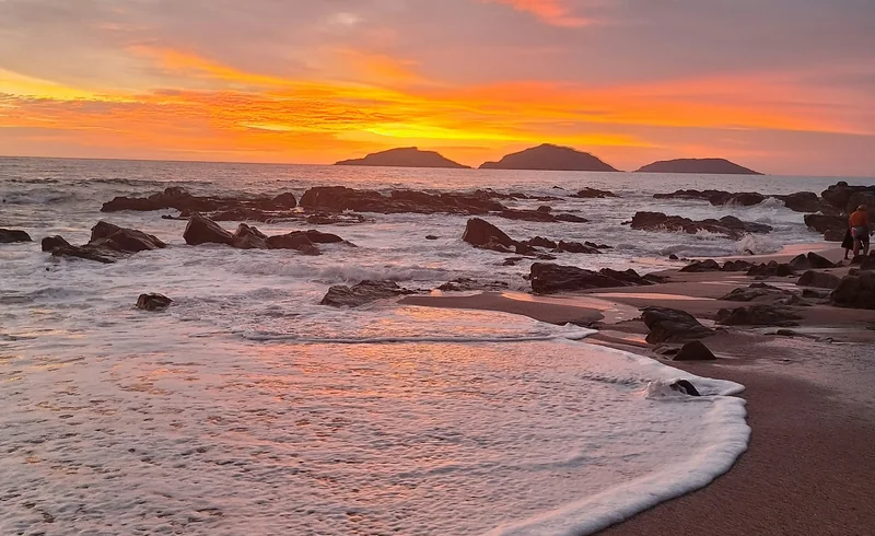 Three islands silhouetted beneath an orange and pink sunset, with waves and rocks along the Mazatlán shoreline.