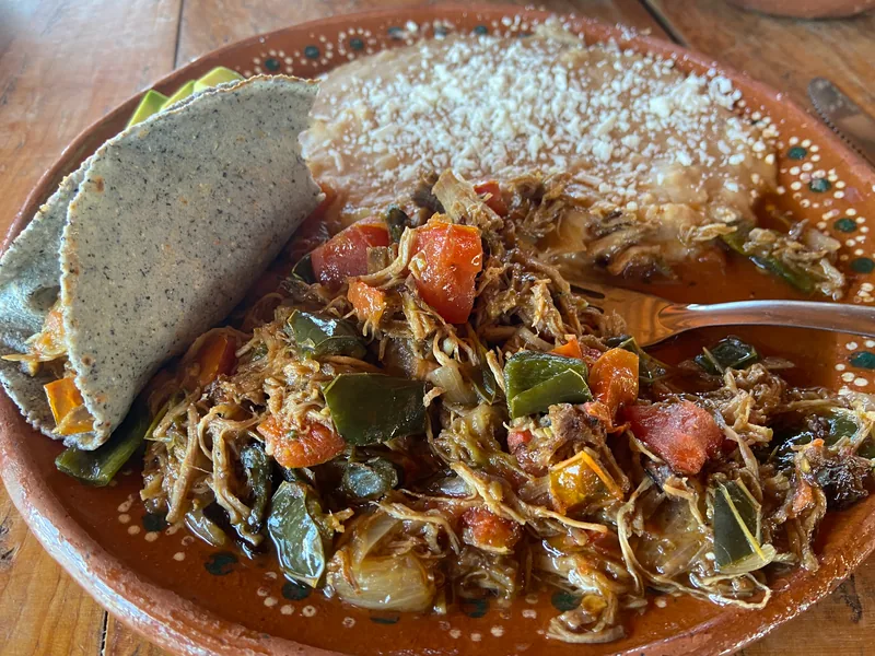 A traditional Mexican dish featuring a blue corn tortilla topped with shredded meat, beans, and fresh garnishes.